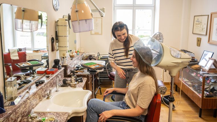 Un salon de coiffure avec un client assis sur un fauteuil de coiffure. La coiffeuse lui parle et un casque est sur la tête du client.