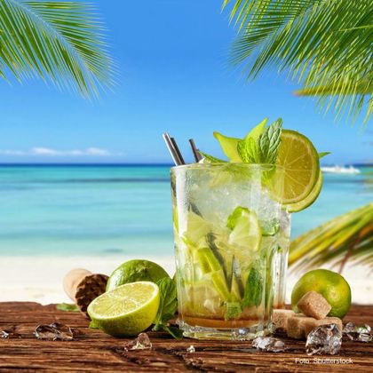 Ein erfrischendes Mojito-Getränk mit Limetten und Minze auf einem Holztisch. Im Hintergrund sind der Ozean und palmengesäumte Strände zu sehen.