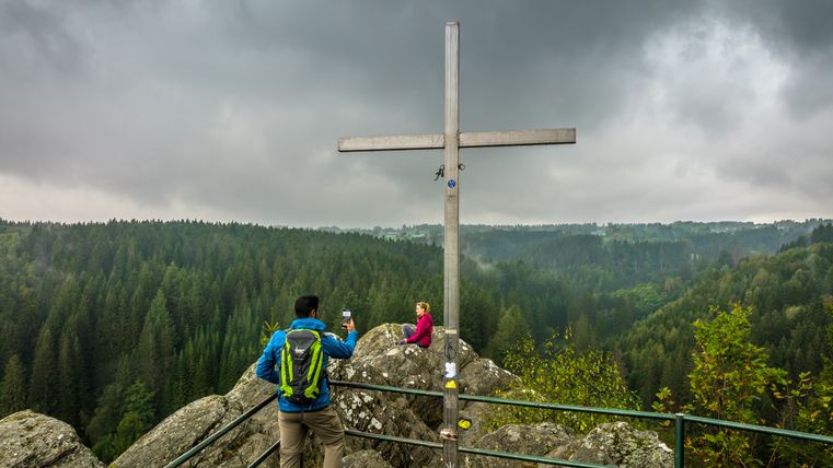 Zwei Personen an einem Aussichtspunkt mit Kreuz, umgeben von Wald und bewölktem Himmel.