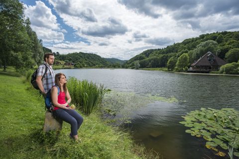 Two people are sitting on the shore of a lake with wooded hills in the background.