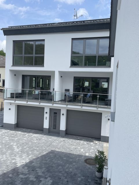 Une maison moderne avec de grandes fenêtres et un balcon. Devant la maison se trouve un parking pavé avec deux garages.