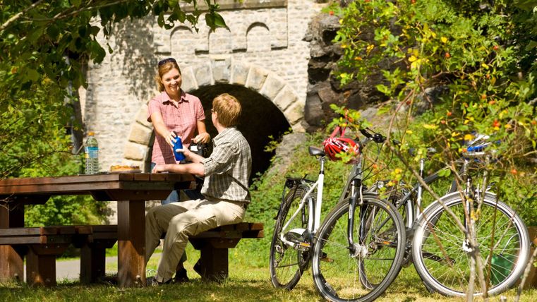 Deux personnes font une pause à une table de pique-nique à côté de vélos, avec un pont de pierre en arrière-plan.