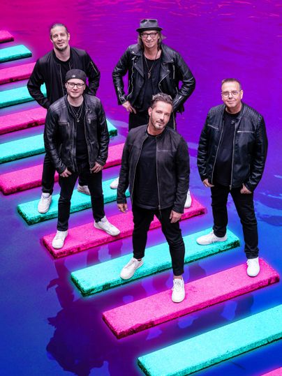 Eine Band steht auf bunten Plattformen über lila Wasser. Alle Mitglieder tragen schwarze Kleidung und sehen stylisch aus.