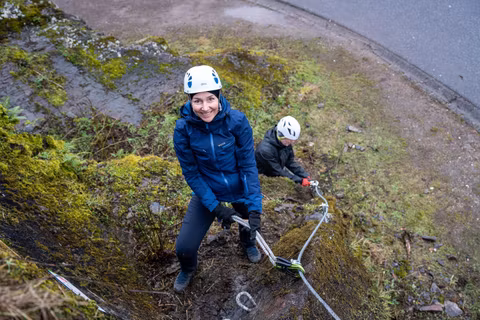 A woman climbs with a rope on a steep, moss-covered wall. In the background, there is another person who is also climbing.