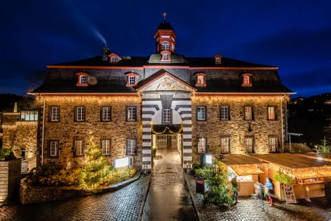 Ein historisches Gebäude mit beleuchteter Fassade in der Dämmerung. Vor dem Gebäude sind Weihnachtsstände und Bäume dekoriert.