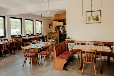 Ein gemütliches Restaurant mit Holzstühlen und Tischen. Große Fenster bieten einen Blick nach draußen und der Raum wirkt einladend und hell.
