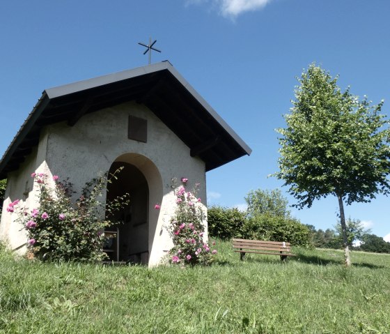 Kleine Kapelle im Grünen, umgeben von blühenden Rosen. Eine Bank steht daneben, ein Baum spendet Schatten. Blauer Himmel im Hintergrund., © Tourist-Information Bitburger Land, Melanie Salzburger