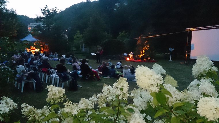 Eine gemütliche Veranstaltung im Freien, bei der Zuschauer im Gras sitzen und einen Film auf einer Leinwand anschauen. Im Vordergrund blühen weiße Blumen, und ein Lagerfeuer sorgt für eine warme Atmosphäre.