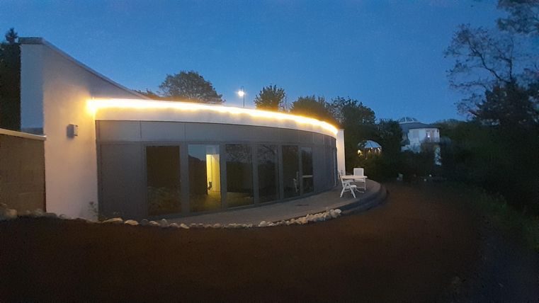 Een modern huis met zachte rondingen straalt zacht in de schemering. Het heeft grote ramen en een goed onderhouden buitenruimte.