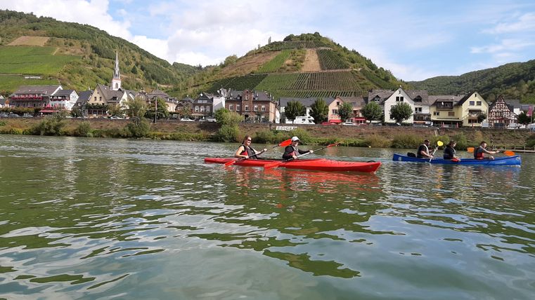 Two kayakers are paddling on a calm river. In the background, picturesque villages and vineyards can be seen.