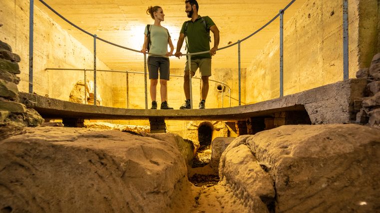 Two people stand on a platform in an ancient Roman well room.