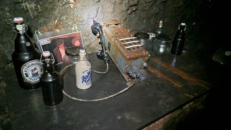 Ein Tisch in einem dunklen Raum mit verschiedenen Flaschen und einem alten Gerät. Im Hintergrund ist ein Foto zu sehen, das auf den Tisch gelehnt ist.