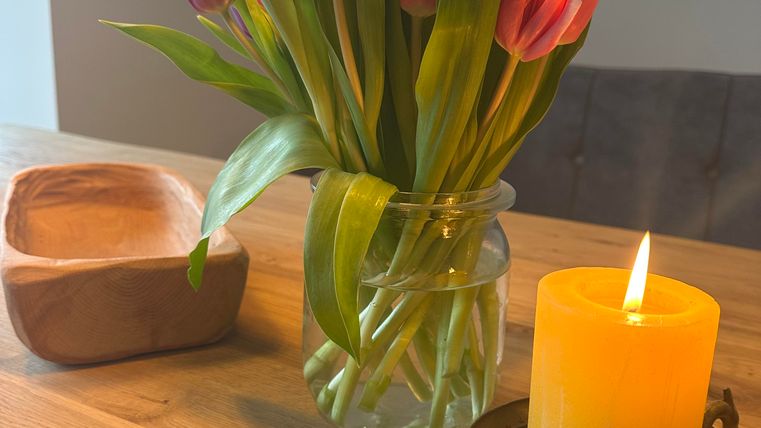 Ein Strauß pinker Tulpen in einem Glasvase steht auf einem Holztisch. Neben der Vase befindet sich eine gelbe Kerze in einem dekorativen Halter.
