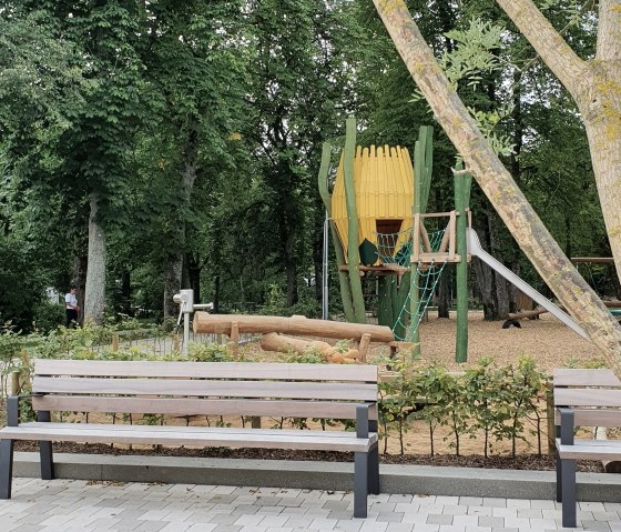 Spielplatz im Park Maximiner Wäldchen in Bitburg mit Bänken, Klettergerüst und Rutsche, umgeben von Bäumen und Grünflächen., © TI Bitburger Land