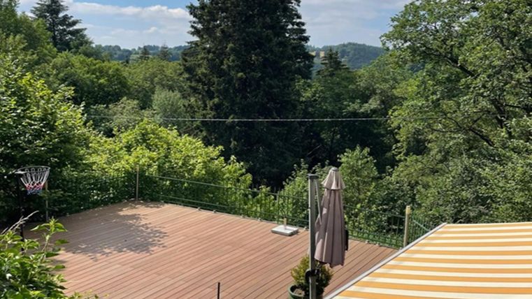 A quiet terrace surrounded by lush greenery and trees. In the background, a blue sky with a few clouds can be seen.