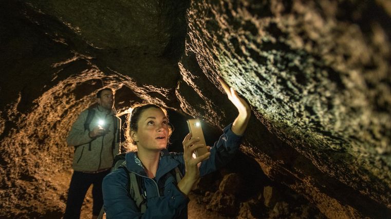 Two people in a cave, illuminating the walls with flashlights. One person holds a smartphone, the other a flashlight.