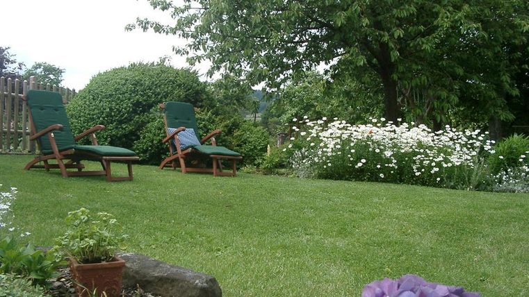 Ein ruhiger Garten mit zwei Liegestühlen und bunten Blumen. Im Hintergrund steht ein großer Baum und das Gras ist gut gepflegt.