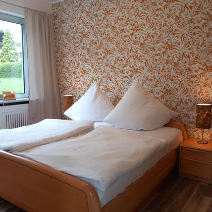 Een gezellig slaapkamer met een tweepersoonsbed en grote kussens. De muren zijn versierd met een bloemmotief en een raam laat daglicht binnen.