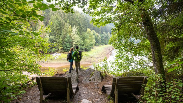 Twee wandelaars staan op een uitkijkpunt dat uitkijkt over een beboste helling en een rivier.