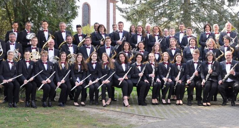 Un grand groupe de musiciens se tient à l'extérieur et joue différents instruments. Tous portent des costumes noirs élégants et sourient à la caméra.