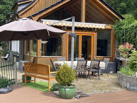 A cozy wooden house with a terrace and a sun umbrella. There are chairs and a table outside to enjoy nature.
