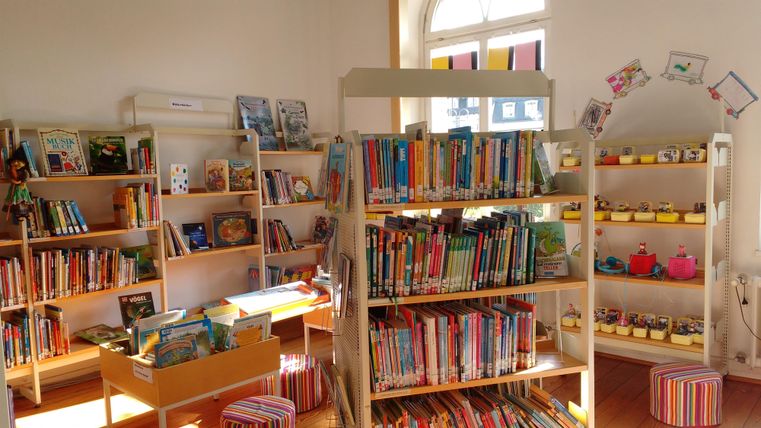 Ein gemütlicher Raum mit Regalen voller Bücher für Kinder. Auf Tischen stehen bunte Spielsachen und kreative Materialien.
