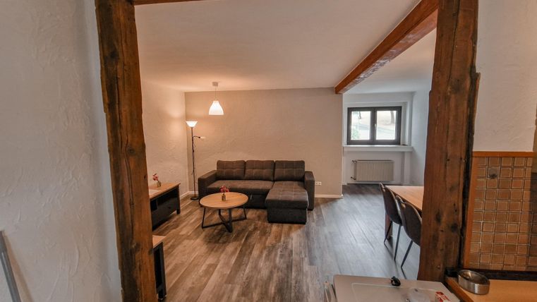 Ein gemütliches Wohnzimmer mit einer grauen Couch und einem Kaffeetisch. Der Raum ist hell und hat Holzbalken sowie eine moderne Beleuchtung.