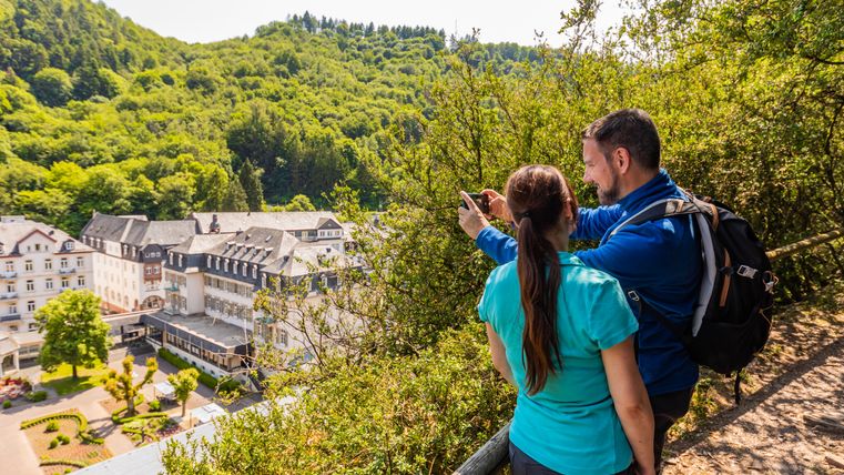 Zwei Personen fotografieren eine Landschaft mit Gebäuden und Wald im Hintergrund.