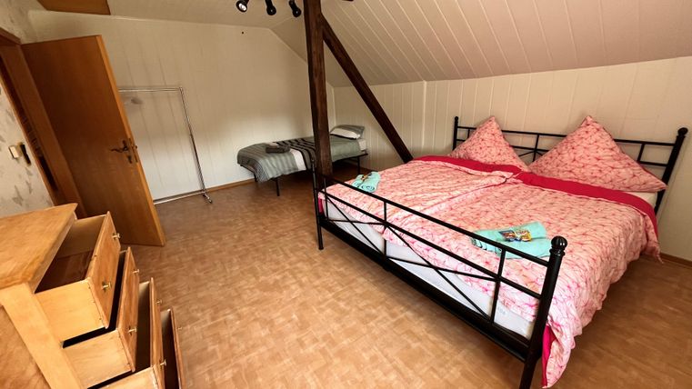 A cozy bedroom with a double bed and pink bedding. Next to the bed, there is a dresser, and there is an additional sleeping space in the corner.