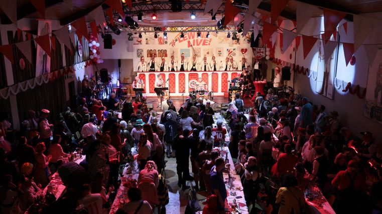 A lively event with many guests in a festively decorated hall. Colorful flags hang from the ceiling and a stage is visible in the background.