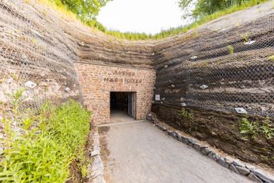 Ein Eingang aus Stein führt in einen Tunnel. Der Weg dorthin ist mit Kies bedeckt. Links und rechts stehen hohe Steinmauern. Oben wächst grünes Gras.