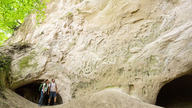 Twee mensen staan voor grote rotsen met grotopeningen in het bos.
