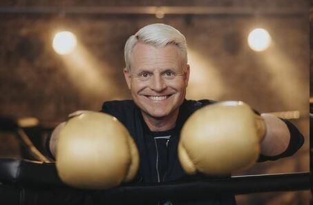 Ein lächelnder Mann mit blonden Haaren trägt goldene Boxhandschuhe. Er steht in einem Boxring und wirkt sportlich und energiegeladen.