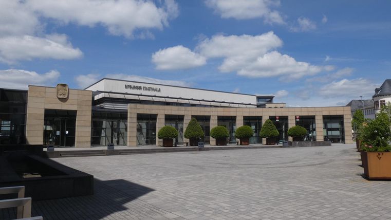 Ein modernes Gebäude mit großen Fenstern und einem Platz davor. Der Himmel ist klar mit einigen Wolken.