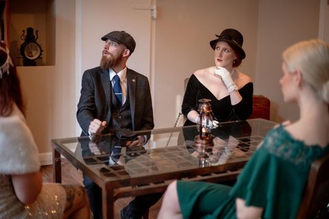 Un groupe de personnes élégamment habillées est assis à une table dans une pièce chic. Les hommes portent des costumes et des chapeaux, tandis que les femmes se présentent en robe de soirée classique.