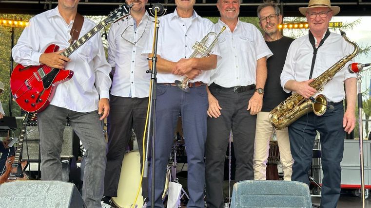 Een band staat op een podium en poseert voor een foto. Ze dragen witte shirts en houden muziekinstrumenten vast.