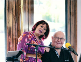 Un couple heureux se tient devant une fenêtre et sourit à la caméra. En arrière-plan, des microphones et une belle vue sont visibles.