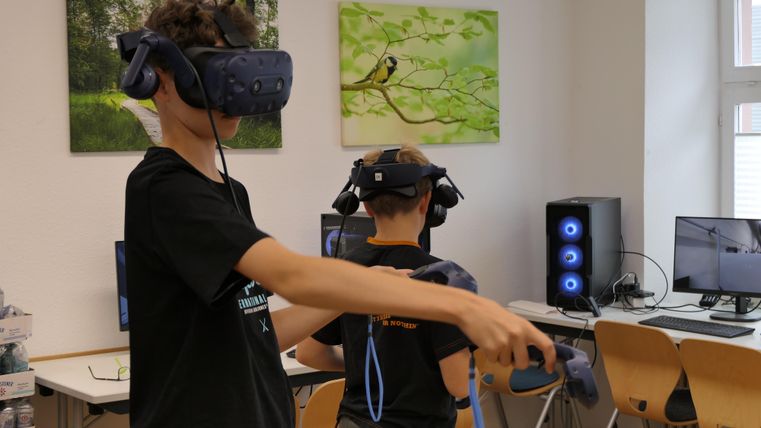 Twee kinderen dragen virtual reality-headsets en interageren met de technologie. Op de achtergrond zijn computers en afbeeldingen aan de muur zichtbaar.