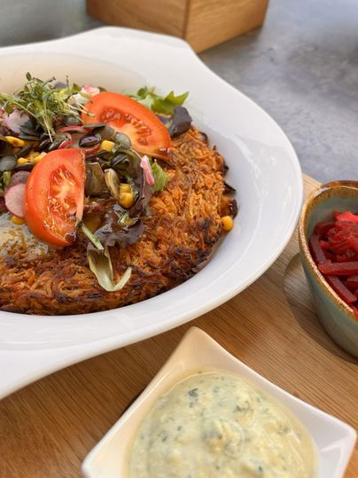 A hearty dish made from grated potatoes, garnished with fresh vegetables and tomato slices. Next to it are two different dips in bowls.