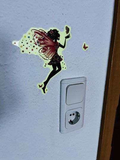 Een mooie wandstickers van een fee met kleurrijke vleugels en vlinders. De sticker bevindt zich naast een stopcontact aan de muur.