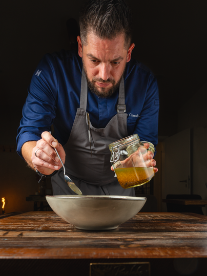 Un cuisinier prépare un repas et ajoute une sauce d'un pot dans un bol. Il se concentre sur la préparation et se tient à une table en bois.