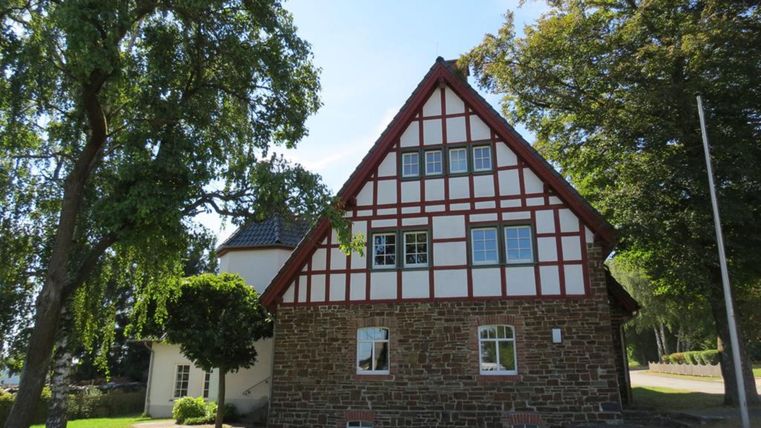 Ein traditionelles Fachwerkhaus mit rotem Gebälk. Umgeben von Bäumen und einer grünen Wiese.