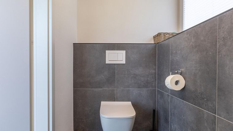 Een modern badkamer met grijze tegels en een eenvoudige toilet. Er is een toiletrolhouder en daglicht dat door een raam valt.