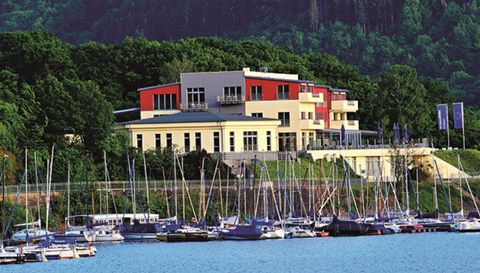 A modern building by the water with many sailboats in the foreground. The surroundings are green and offer a beautiful view of nature.