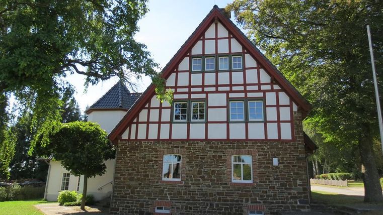 Ein traditionelles Fachwerkhaus mit rotem Dach und weißen Fenstern. Es steht in einer grünen Umgebung mit Bäumen und Rasen.