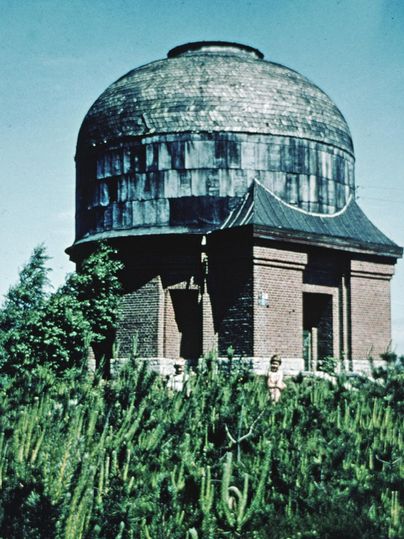 Un observatoire historique avec un dôme en forme d'arc se dresse devant un paysage verdoyant. Autour du bâtiment, différentes plantes poussent.