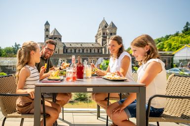 Familie mit zwei Kindern sitzt bei sonnigem Wetter auf der Terrasse einer Gaststätte und genießt ein gemeinsames Essen. Im Hintergrund die eindrucksvolle romanische Basilika Maria Laach mit ihren markanten Türmen. Auf dem Tisch stehen bunte Getränke und verschiedene Speisen. Die Stimmung ist fröhlich und entspannt.