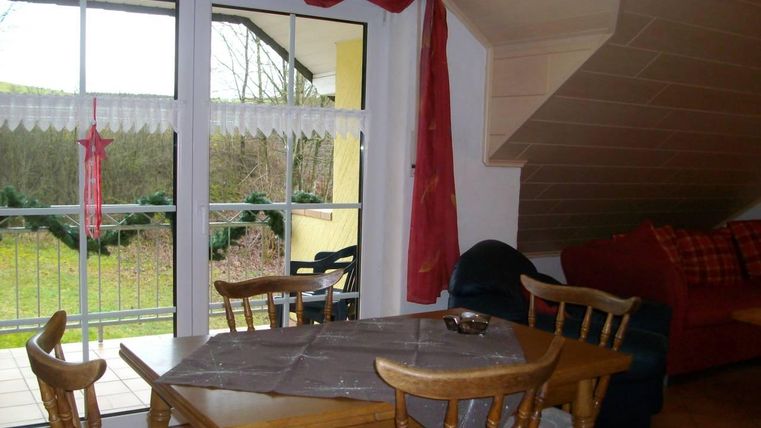 Een gezellige eetruimte met een houten tafel en stoelen. Op de achtergrond zijn grote ramen die uitkijken op een groen landschap.