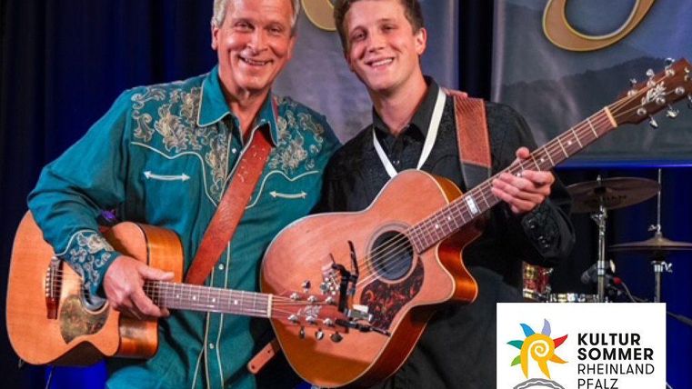 Two musicians are standing on stage and smiling at the camera. Both are holding guitars in their hands.