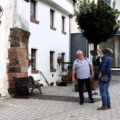 Twee mannen staan voor een historisch gebouw met een oude boog. De een wijst naar iets terwijl de ander luistert.